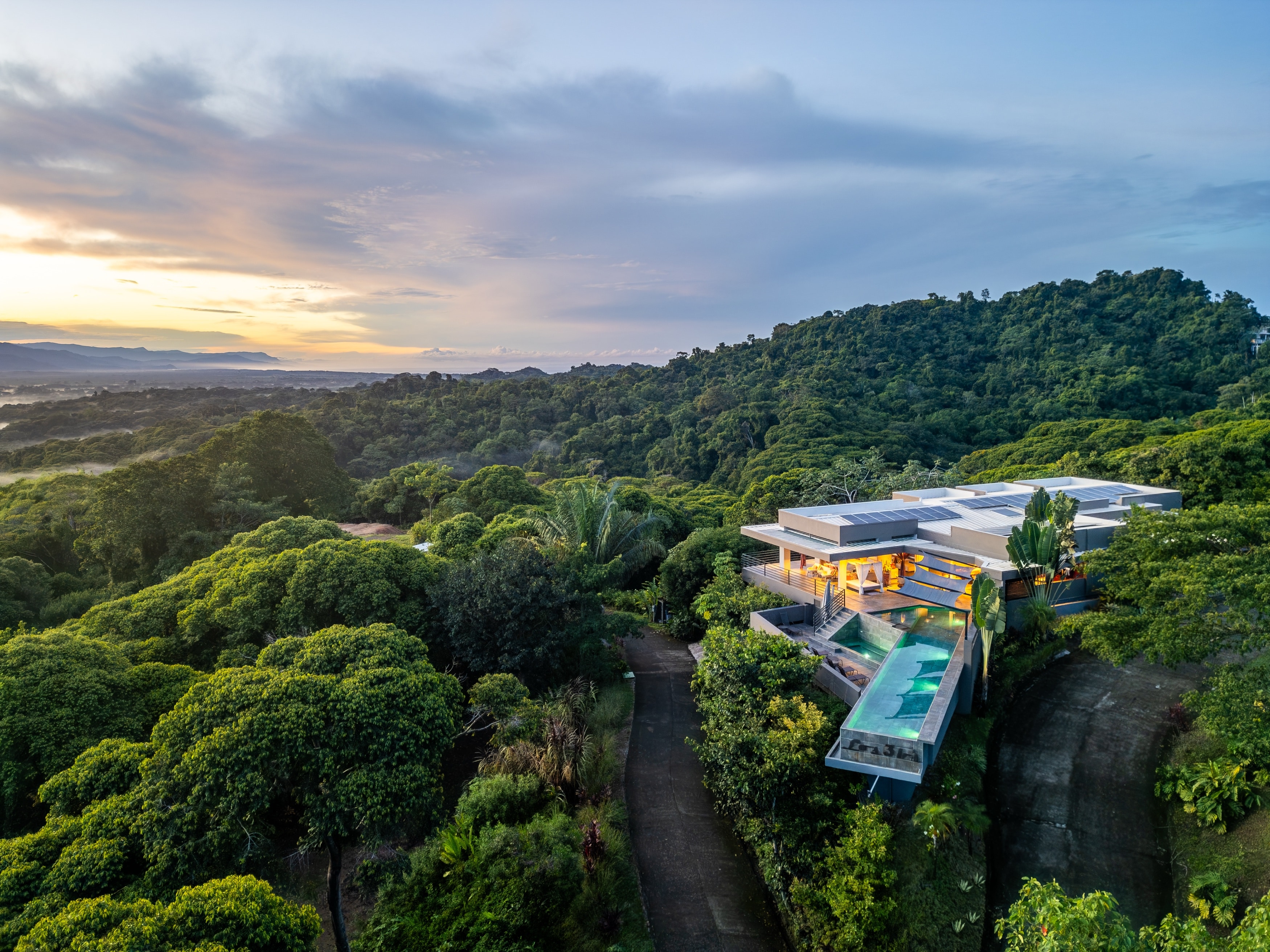 Aerial at sunset — the villa on its ridge above the canopy, the coast beyond